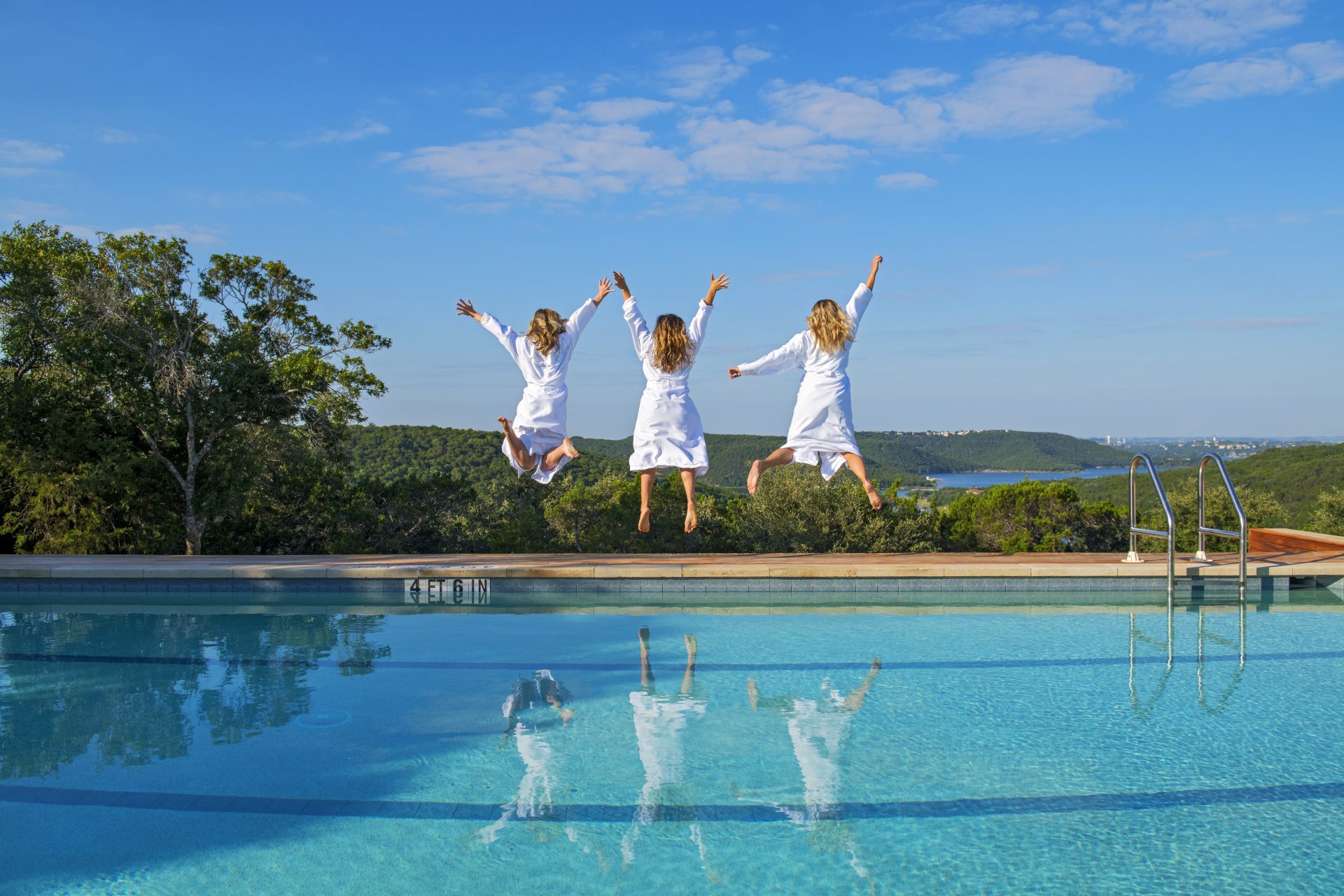 Three women in robes jumping on pool deck with arms up looking at mountain landscape