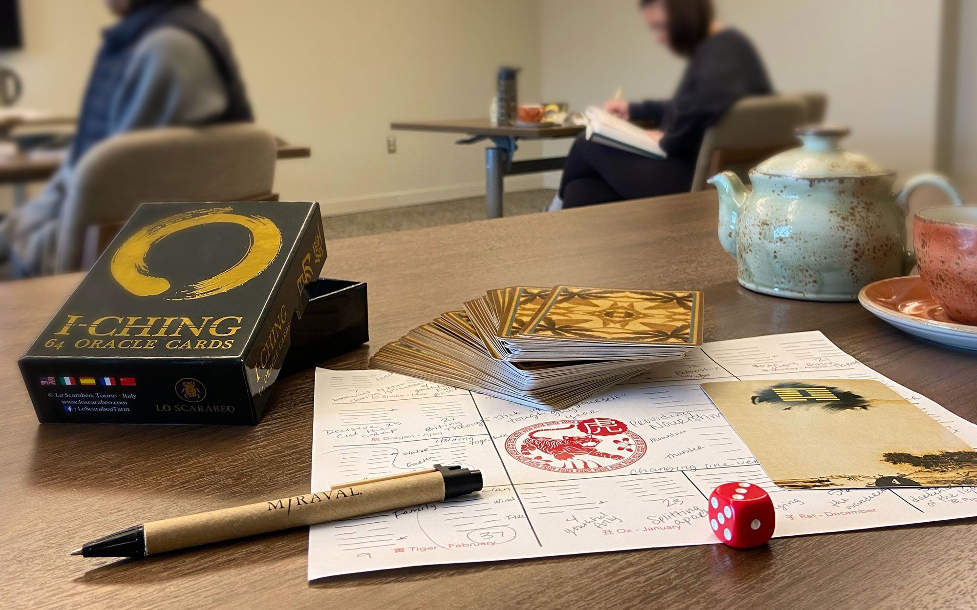 Table with oracle cards, a dice, miraval pen and tea pot, people in the background