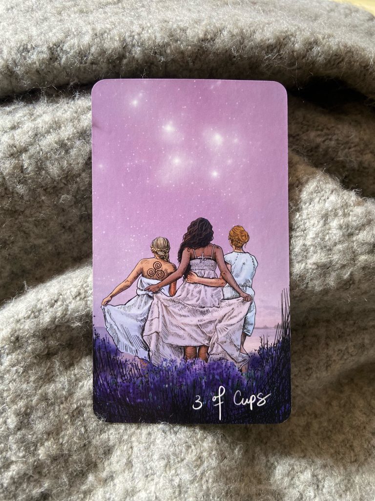 Tarot card, three of cups with three women arm in arm in front of purple star sky